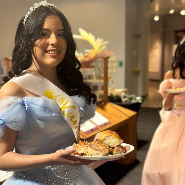 Smiling woman in a light blue gown and tiara holding a plate of food, including a roll and meat.
