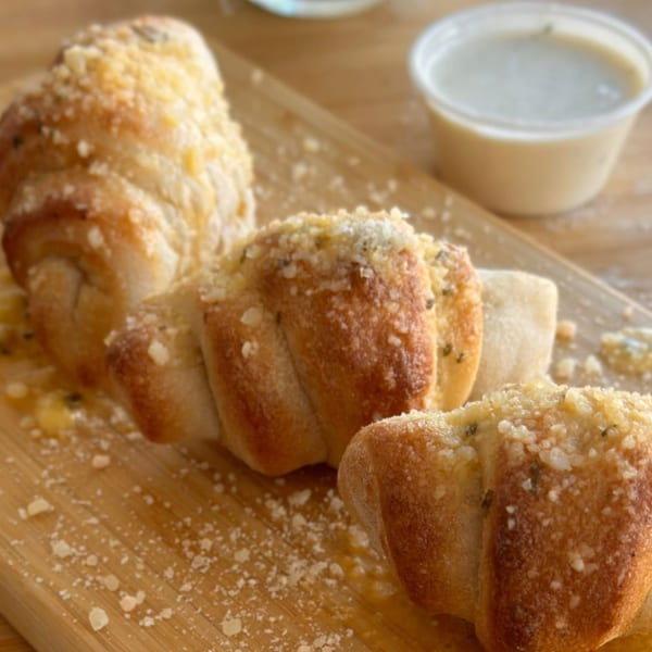 Three garlic knots, sprinkled with parmesan cheese, on a wooden board with dipping sauce.