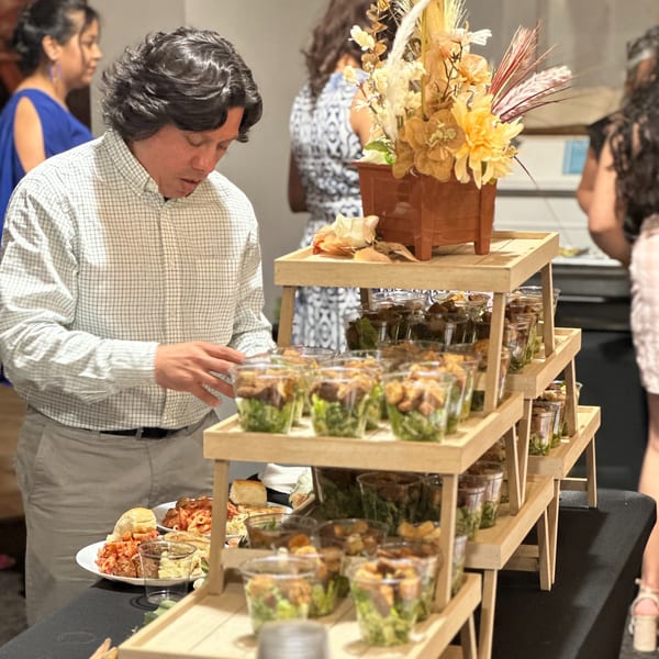 A man in a plaid shirt serves himself pasta from a buffet with many individual salad cups.