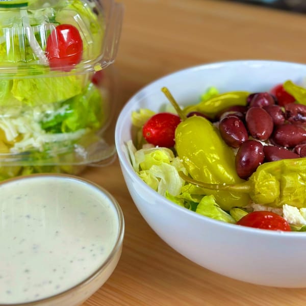 A fresh salad with lettuce, tomatoes, olives, and peppers, with a side of dressing.