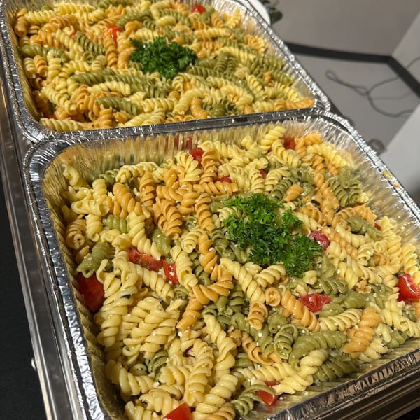 Two foil trays filled with colorful rotini pasta salad, cherry tomatoes, and parsley garnish.